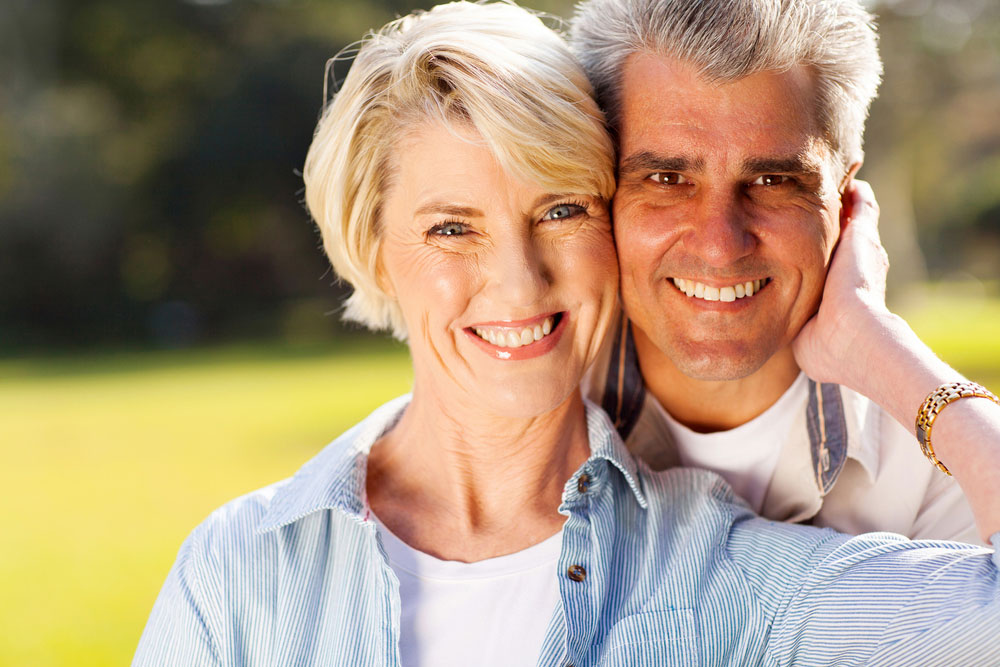 Middle-aged couple smiling outdoors in casual clothing, green lawn background.