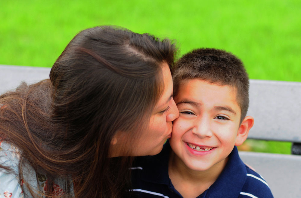 Mother kissing young son's cheek, both smiling against green and white background.