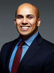 Professional headshot of smiling man in dark blazer and blue shirt against gray background.