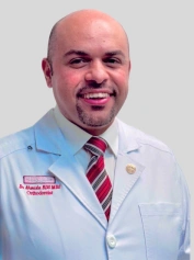 Professional male dentist in white coat and striped tie smiling at camera in clinical setting.