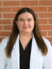 Female dental professional in white coat smiling at camera against brick wall background.