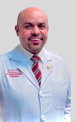 Professional male dentist in white coat and striped tie smiling at camera in dental office setting.